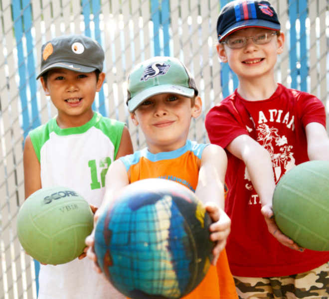 Three young kids stand together holding volleyballs out towards the photographer.